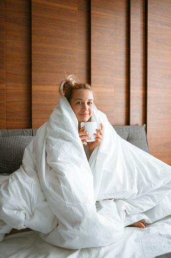 Cute smiling teen girl under blanket in white bed in bedroom woken up and drink tea. Breakfast in bed, lazy morning. High quality photo