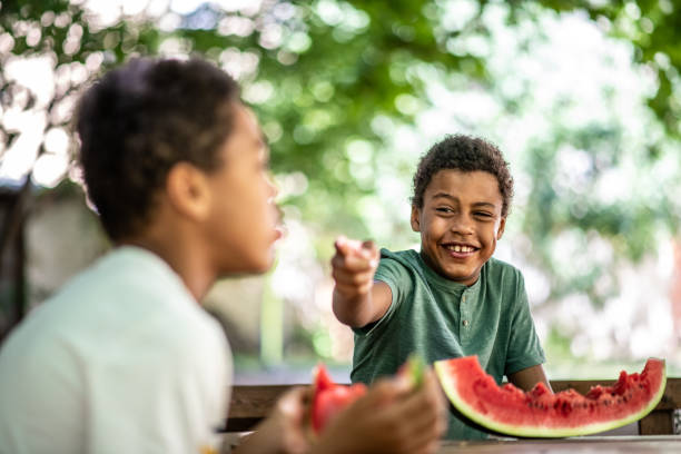 istock Two boys are sitting at a table eating a watermelon, talking, one is laughing and pointing something with his finger 1263348354