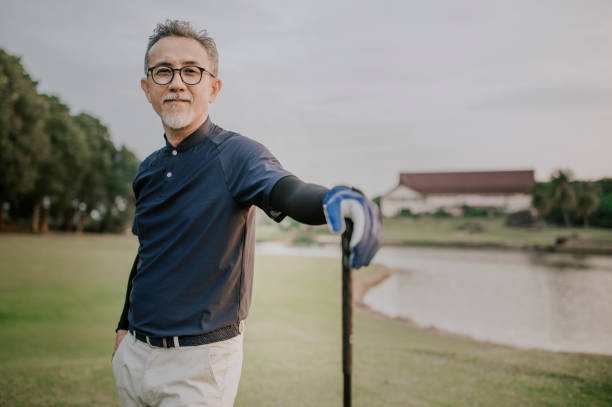 アジアの中国人の現役の先輩ゴルファーは、ゴルフの試合を待って見ている後、ゴルフコースで彼のゴルフクラブのドライバーと一緒に立っています - attractive male confidence satisfaction happiness ストックフォトと画像