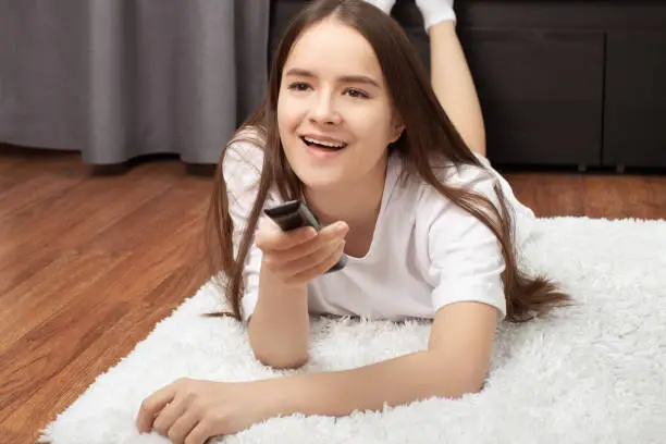 Photo of Exciting TV, movies, TV shows. The girl lies at home with a remote control in her hands, watches TV, joyful, surprised