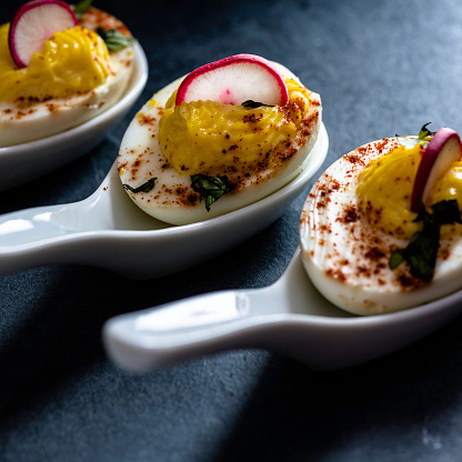 Deviled eggs garnished with smoked spanish paprika, sliced radishes, and chopped fresh basil; served on individual ceramic spoons.