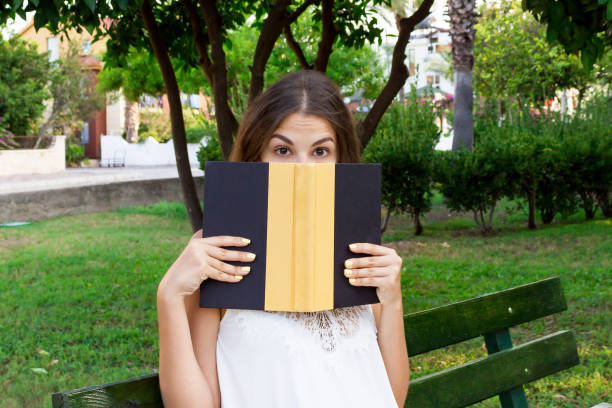 a jovem bonita está cobrindo o rosto com um livro por causa da surpresa e do choque. mulher engraçada emocional está segurando um livro em suas mãos no parque - question mark obscured face asking women - fotografias e filmes do acervo