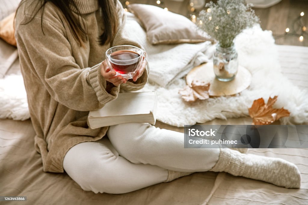 Cozy autumn or winter at home, a woman in a knitted sweater Cozy autumn or winter at home, a woman in a knitted sweater with a Cup of tea and a book . Hygge Stock Photo