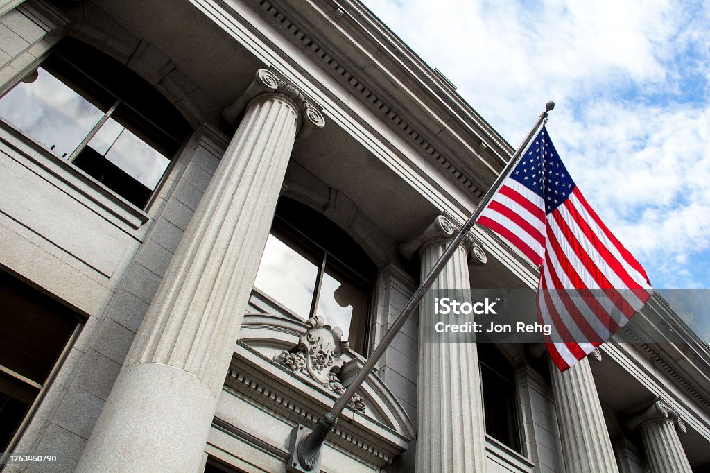 American flag flying over government building in city, blue sky and clouds Stone granite official government building in city with American flag waving and flying in the wind, looking up, outside, judicial, freedom, civil courts, columns, criminal justice, law Government Stock Photo