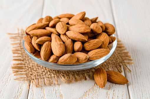 Tasty raw almonds in a glass saucer on a white wood table. Unroasted nuts as antioxidant and protein source for ketogenic diet and vegetarianism. Healthy eating. Front view.