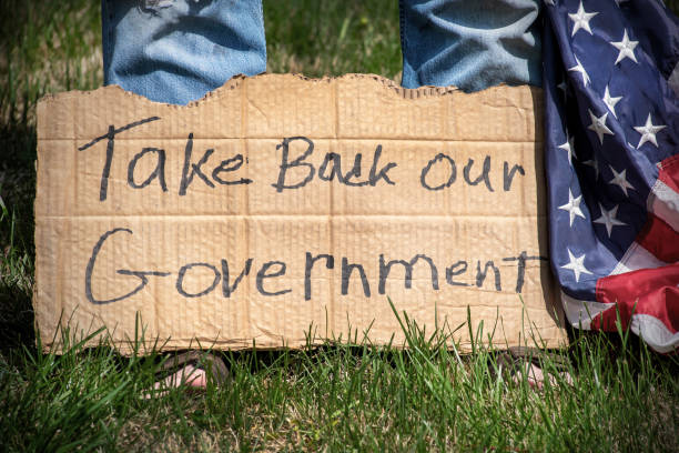 Closeup man standing in yard with cardboard sign Take Back Our Government at his feet, and American flag Man standing on lawn with American flag and holding handmade hand written cardboard sign Take Back Our Government, protesting, rebellion, anti-government usa flag shoes stock pictures, royalty-free photos & images