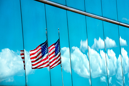 American flag rippling, moving, waving in the wind, reflected in glass window, with blue sky and white clouds, freedom, independence, liberty