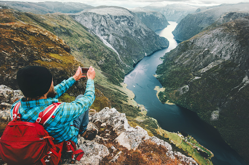 Man backpacker taking photo by smartphone relaxing on mountain cliff traveling in Norway adventure lifestyle active vacations modern technology connection concept