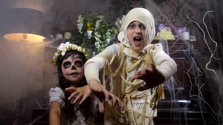 Two girls dressed in Halloween mummy and Santa Muerte costumes  posing and scaring asking for treat in old dark Gothic interior covered with cobwebs and mysterious fog. 4K slow motion 50 fps