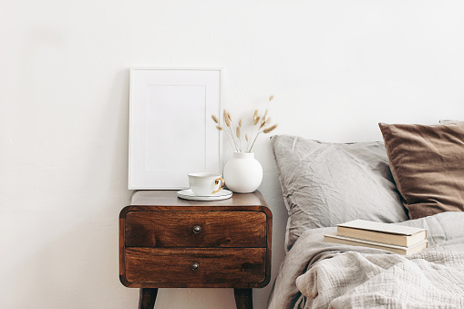 Portrait white frame mockup on retro wooden bedside table. Modern white ceramic vase with dry Lagurus ovatus grass and cup of coffee. Beige linen and velvet pillows in bedroom, Scandinavian interior.