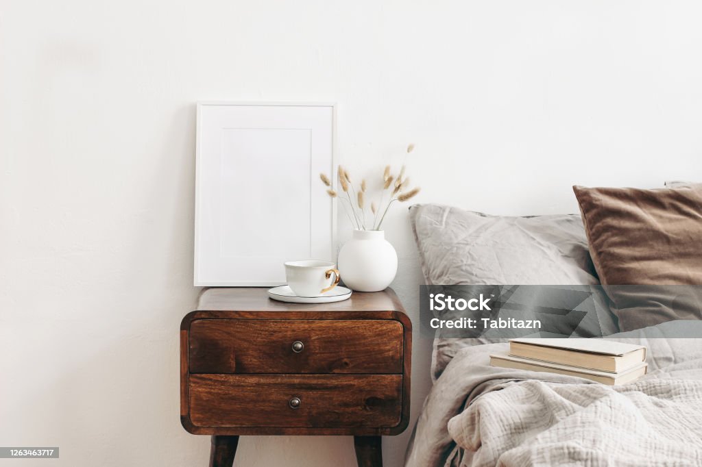 Portrait white frame mockup on retro wooden bedside table. Modern white ceramic vase with dry Lagurus ovatus grass and cup of coffee. Beige linen and velvet pillows in bedroom. Scandinavian interior. Portrait white frame mockup on retro wooden bedside table. Modern white ceramic vase with dry Lagurus ovatus grass and cup of coffee. Beige linen and velvet pillows in bedroom, Scandinavian interior. Night Table Stock Photo