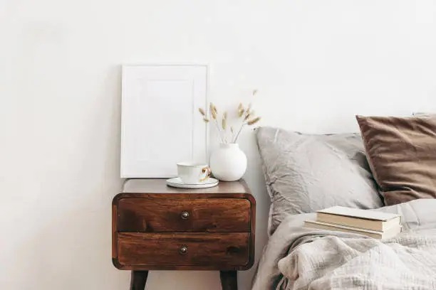 Photo of Portrait white frame mockup on retro wooden bedside table. Modern white ceramic vase with dry Lagurus ovatus grass and cup of coffee. Beige linen and velvet pillows in bedroom. Scandinavian interior.