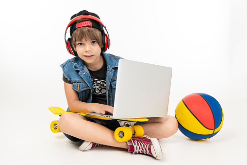 european boy sitting on the floor with laptop, skateboard and basketball on a white background