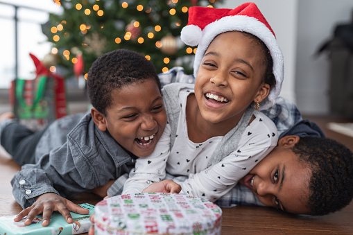Three (3) siblings of African American descent enjoy Christmas Day together. They are laughing and laying on top of each other.