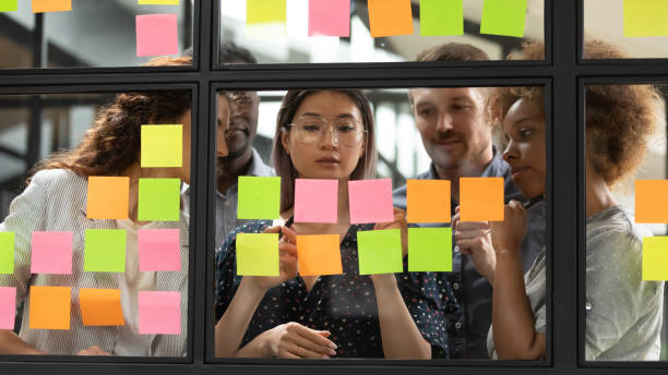 Diverse businesspeople discussing, planning project on sticky papers together Diverse businesspeople discussing, planning new project on sticky papers on glass board in office together, creative employees team sharing startup ideas, writing tasks on stickers at briefing scrum stock pictures, royalty-free photos & images