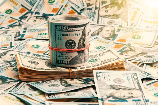A background of scattered one-hundred-dollar bills over the entire space, close-up in the middle of a stack and a roll of dollars in the shape of a ship. Top and side view. Color and contrast images.