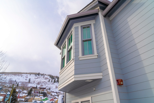 Home exterior in Park City Utah with bay window and gray horizontal wall siding. Colorful homes on a snowy hill against cloudy winter sky can be seen in the background.