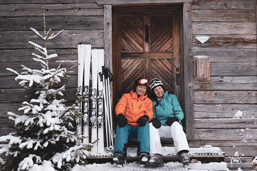 happy fit senior couple in their seventies sitting at wooden mountain hut in ski clothes with skiing helmets and skis on winter vacation day