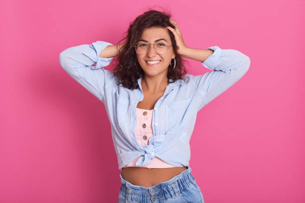 istock Happy woman with hands on head looking directly at camera with charming smile, expressing positive emotions, stands against rosy wall, wearing jeans and blue shirt. 1263546360