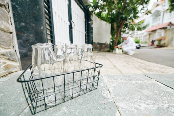 Wire crate with empty glass bottles and milkman putting bottles with fresh milk in front of customers entrance door in background