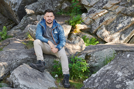 Portrait of mature man in casual clothing sitting on rocks and looking at camera during hiking