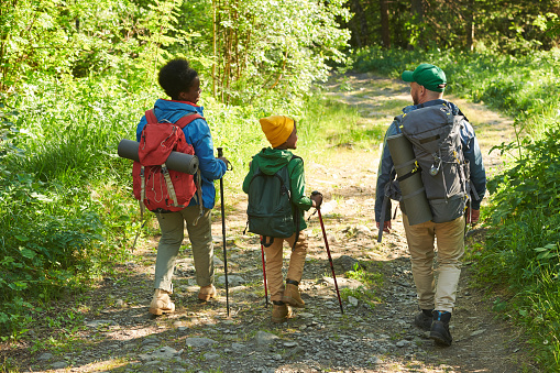 A mother and her daughter take a hike on a warm summers evening as they stay fit together.  They are both dressed casually, are carrying backpacks and have walking sticks in hand.