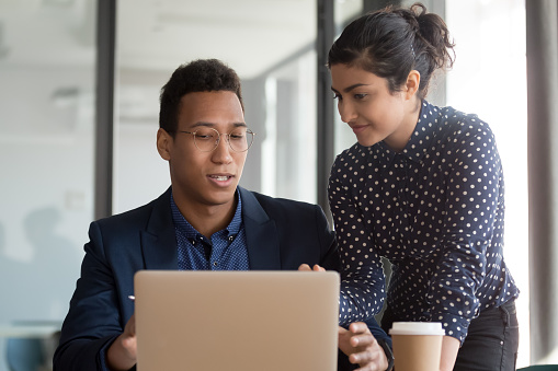 Young diverse black male and indian female coworkers working together on start up project. African american businessman discussing contract details or software with smiling colleague intern at office.