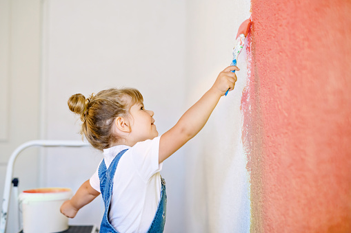 Funny little toddler girl painting the wall with color in new house. Family repair apartment home. Happy baby child paints the wall, choosing color with palette, having fun with brush, indoors.