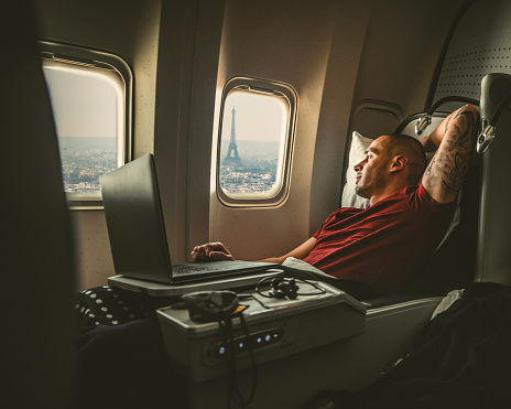 Casually dressed middle aged man working on laptop in aircraft cabin during his business travel, while looking through window at  landmark Eiffel tower.
