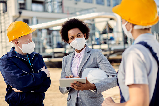 Black businesswoman and two construction workers wearing protective face masks while talking about ongoing project outdoors.