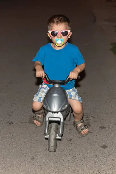 Photo of a boy with funny sunglasses and a pacifier rides a children's motorcycle