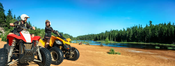 quad atv alla guida delle persone. felice coppia sorridente ciclisti sulla spiaggia nel bellissimo lago. foy, foyross lake, sudbury, ontario, canada. - off road vehicle quadbike 4x4 adventure foto e immagini stock