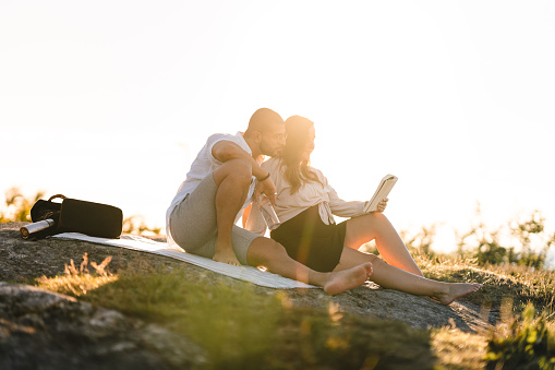 A couple in love, sitting close together on a picnic blanket in the sun. They are reading a book together.