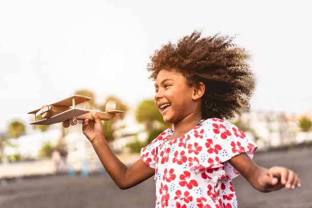 Photo of African kid running on the beach while playing with wood toy airplane at sunset, Travel and youth lifestyle concept - Main focus on hand holding plane