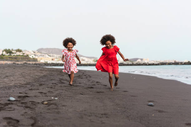 Afro twins sisters running on the beach together - Youth lifestyle and travel concept - Main focus on right kid face Afro twins sisters running on the beach together - Youth lifestyle and travel concept - Main focus on right kid face colombian family stock pictures, royalty-free photos & images