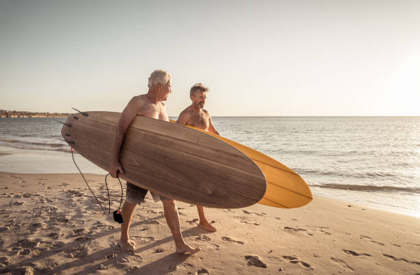 deux hommes mûrs marchant avec des planches de surf sur la belle plage appréciant le paradis et le mode de vie de retraite. attrayant ajustement amis adultes âgés ayant du plaisir à surfer. chez de vraies personnes actives et en bonne santé. - senior adult surfing aging process sport photos et images de collection