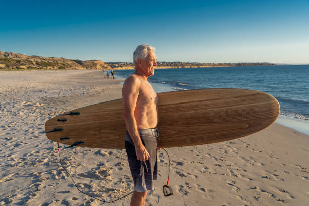 surfeur australien mûr attrayant d’homme avec la planche de surf vintage fraîche sur la plage au coucher du soleil. adulte aîné heureux d’être de retour pour surfer . aventure sportive en plein air, personnes âgées actives et mode de vie à la r - senior adult surfing aging process sport photos et images de collection