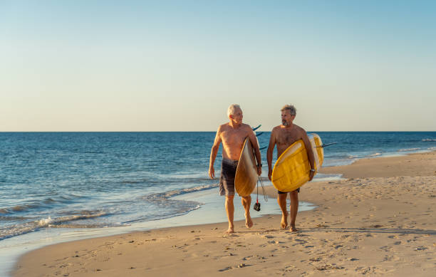 deux hommes mûrs marchant avec des planches de surf sur la belle plage appréciant le paradis et le mode de vie de retraite. attrayant ajustement amis adultes âgés ayant du plaisir à surfer. chez de vraies personnes actives et en bonne santé. - senior adult surfing aging process sport photos et images de collection