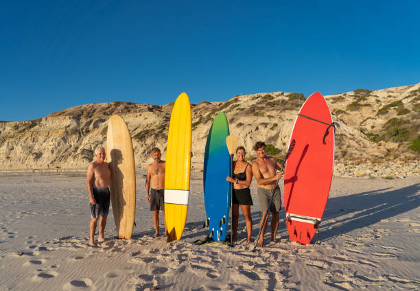 gruppo di surfisti, donne e uomini senior, che tengono le loro tavole da surf colorate sulla spiaggia remota. amici maturi in pensione che si godono il surf e lo stile di vita all'aperto in persone sane e invecchiano nel mondo moderno. - senior adult surfing aging process sport foto e immagini stock