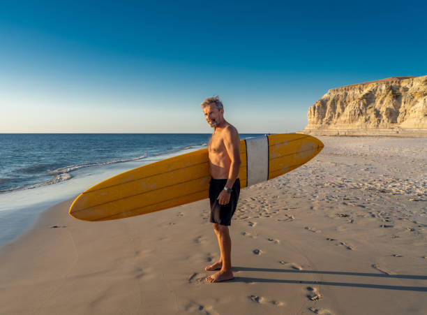 surfeur australien mûr attrayant d’homme avec la planche de surf vintage fraîche sur la plage au coucher du soleil. adulte aîné heureux d’être de retour pour surfer . aventure sportive en plein air, personnes âgées actives et mode de vie à la r - senior adult surfing aging process sport photos et images de collection