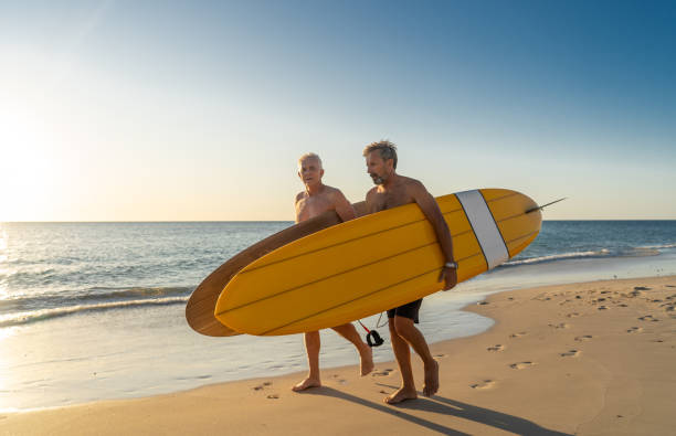 due uomini maturi che camminano con tavole da surf sulla bellissima spiaggia godendo del paradiso e dello stile di vita pensionistico. attraente vestibilità amici adulti anziani che si divertono a navigare. in persone reali attive e sane. - senior adult surfing aging process sport foto e immagini stock