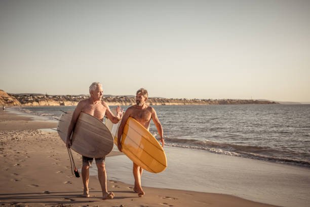 deux hommes mûrs marchant avec des planches de surf sur la belle plage appréciant le paradis et le mode de vie de retraite. attrayant ajustement amis adultes âgés ayant du plaisir à surfer. chez de vraies personnes actives et en bonne santé. - senior adult surfing aging process sport photos et images de collection