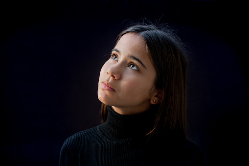 Portrait of a young caucasian girl, wearing a black turtleneck pullover and looking up to the left side. Black background.