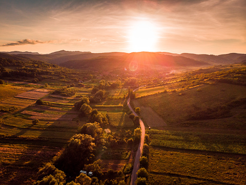 Color image depicting an aerial view of a rural landscape with snow patches in the Transylvania region of Romania. The area is heavily forested with hills in the distance and the orange glow of sunset. A winding road runs directly through the image. Room for copy space.