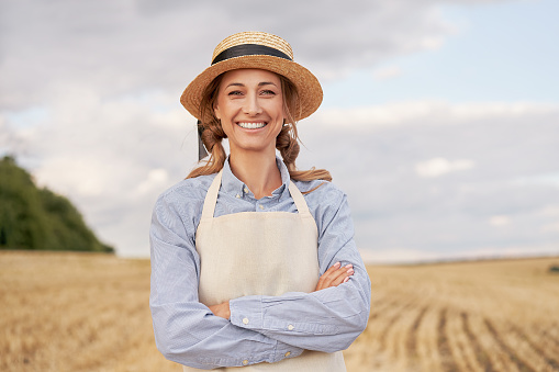 Woman farmer straw hat apron standing farmland smiling Female agronomist specialist farming agribusiness Happy positive caucasian worker agricultural field Girl arms crossed cloudy sky background