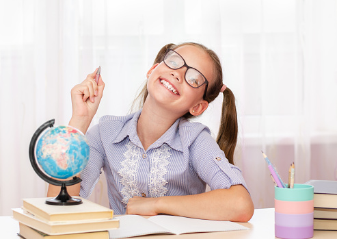 Education and school concept. Smiling happy child in glasses is sitting at the desk and holding the pen . Cute little student girl is studying.