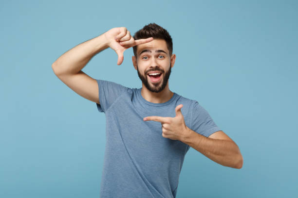 istock Young cheerful funny man in casual clothes posing isolated on blue wall background, studio portrait. People sincere emotions lifestyle concept. Mock up copy space. Making hands photo frame gesture. 1263607260