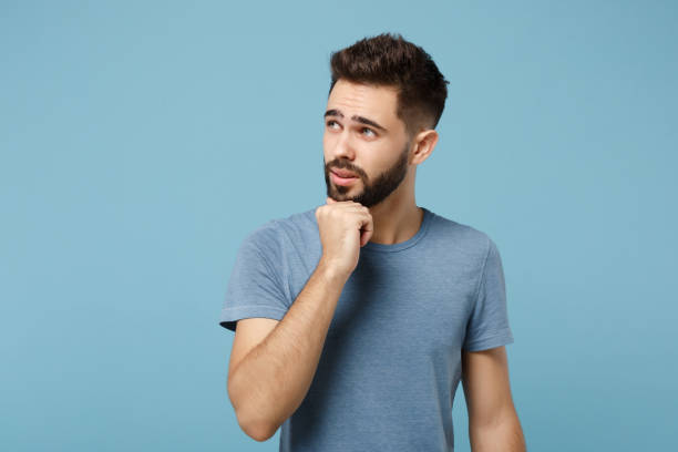 istock Young pensive man in casual clothes posing isolated on blue wall background, studio portrait. People sincere emotions lifestyle concept. Mock up copy space. Put hand prop up on chin, looking aside. 1263607341