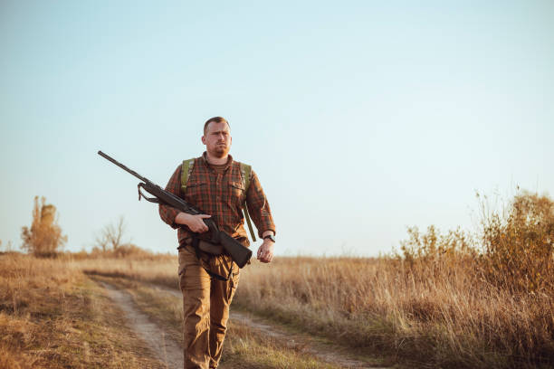 istock Strong young hunter with red beard holding his gun and walking along the dirt road under blue sky 1263607345