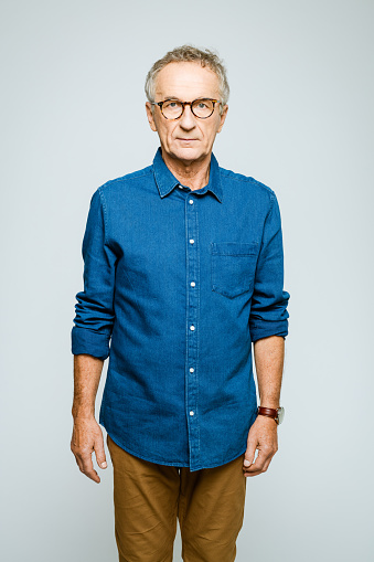 Portrait of elderly man wearing denim shirt and eyeglasses looking at camera. Confident senior entrepreneur, studio shot against grey background.